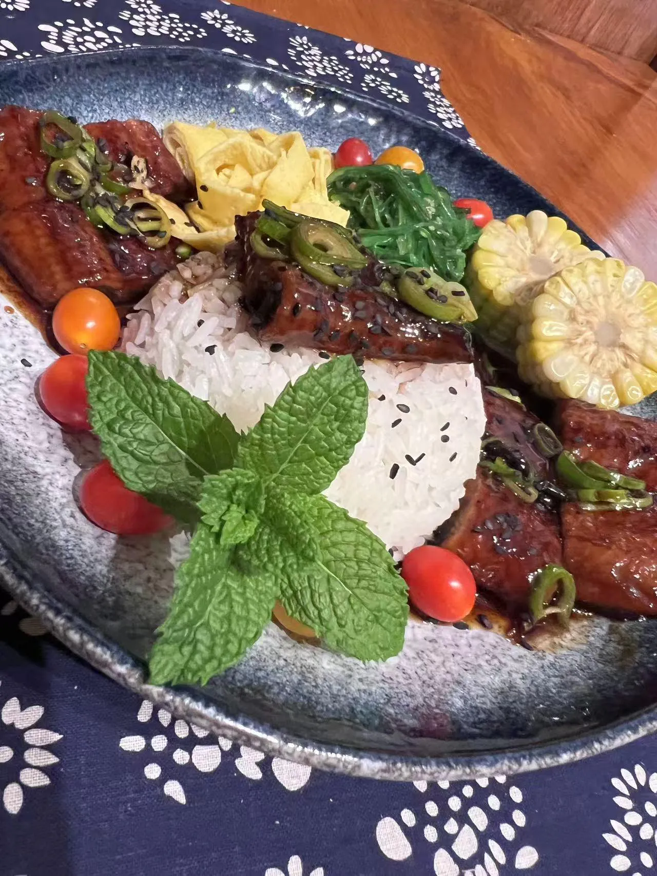 Unagi don rice bowl with glazed eel, corn, cherry tomatoes and mint garnish on blue floral tablecloth at Endless Art And Food, an Asian Fushion Restaurant in Catskill