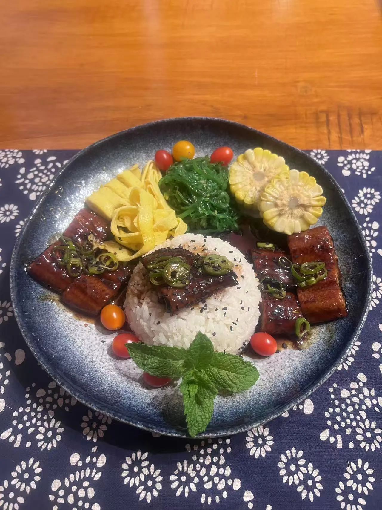 Unagi don rice bowl topped with grilled eel, tamagoyaki, seaweed salad and corn on blue floral cloth at Endless Art And Food, an Asian Fushion Restaurant in Catskill